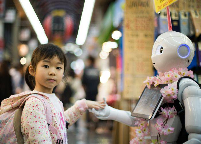 A young girl holding hands with a Pepper robot while looking towards the camera.